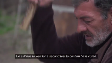 Thumbnail from a video showing a man working with a wooden tool with the subtitle 'He still has to wait for a second test to confirm he is cured.'